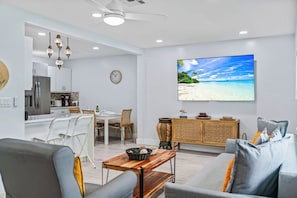 A modern living room with a flat-screen TV displaying a beach scene, a dining area with a clock, and a kitchen with a refrigerator.