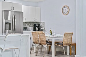 A modern kitchen with white cabinetry, a stainless steel refrigerator, a coffee maker, and a dining table with wicker chairs.