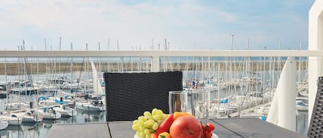 Une table avec une assiette de fruits et un verre d’eau, donnant sur une marina où sont amarrés des bateaux.
