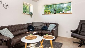 Un soggiorno moderno con un divano angolare in pelle marrone, due tavolini bianchi con sottopiatti intrecciati, un tappeto soffice grigio e un'ampia finestra con vista sul verde.