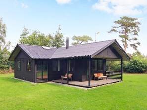 A small, dark-colored cabin with a metal roof, a porch with two chairs, and a chimney.