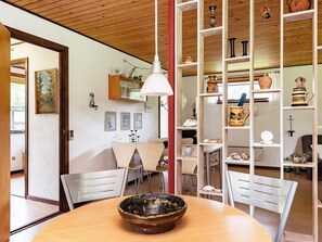 A room with a wooden ceiling, a round table with a bowl, white chairs, and shelves with pottery.