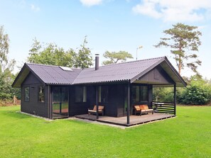 A small, dark-colored cabin with a metal roof, a porch with two wooden benches, and a grassy lawn.