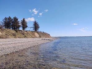A pebbly beach with clear water, trees, and a distant shoreline. - 6 Person Holiday Home in Oster Assels (Øster Assels)