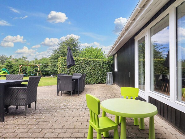 An outdoor patio with a table and chairs, a swing set, and a hedge.