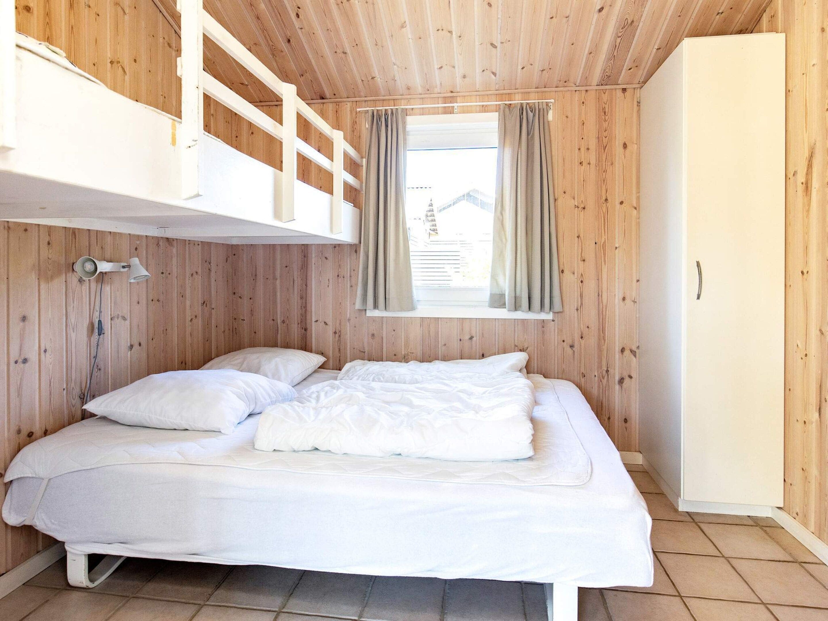 Ein Stockbett mit weißer Matratze und Kissen, ein Fenster mit Vorhängen und eine Holzwand.