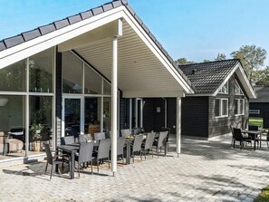 A covered outdoor seating area with tables and chairs, a clear sky, and a house with a dark exterior and a sloped roof. - 5 Star Holiday Home in Vaeggerlose (Vaeggerlose)