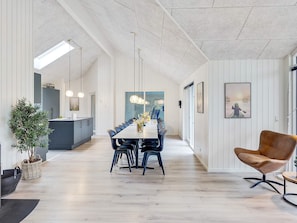 A modern dining area with a long table, chairs, pendant lights, and a view of the kitchen.