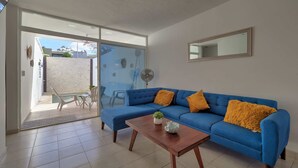A modern living room with a blue sofa, wooden coffee table, and a sliding glass door leading to an outdoor area. - Casita el Pinar - Yucatan Home Rentals (Mérida)