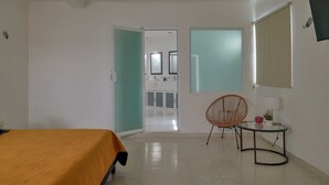 A hotel room with a bed, a chair, a small table, a TV, and a bathroom visible through an open door. - Casita el Pinar - Yucatan Home Rentals (Mérida)
