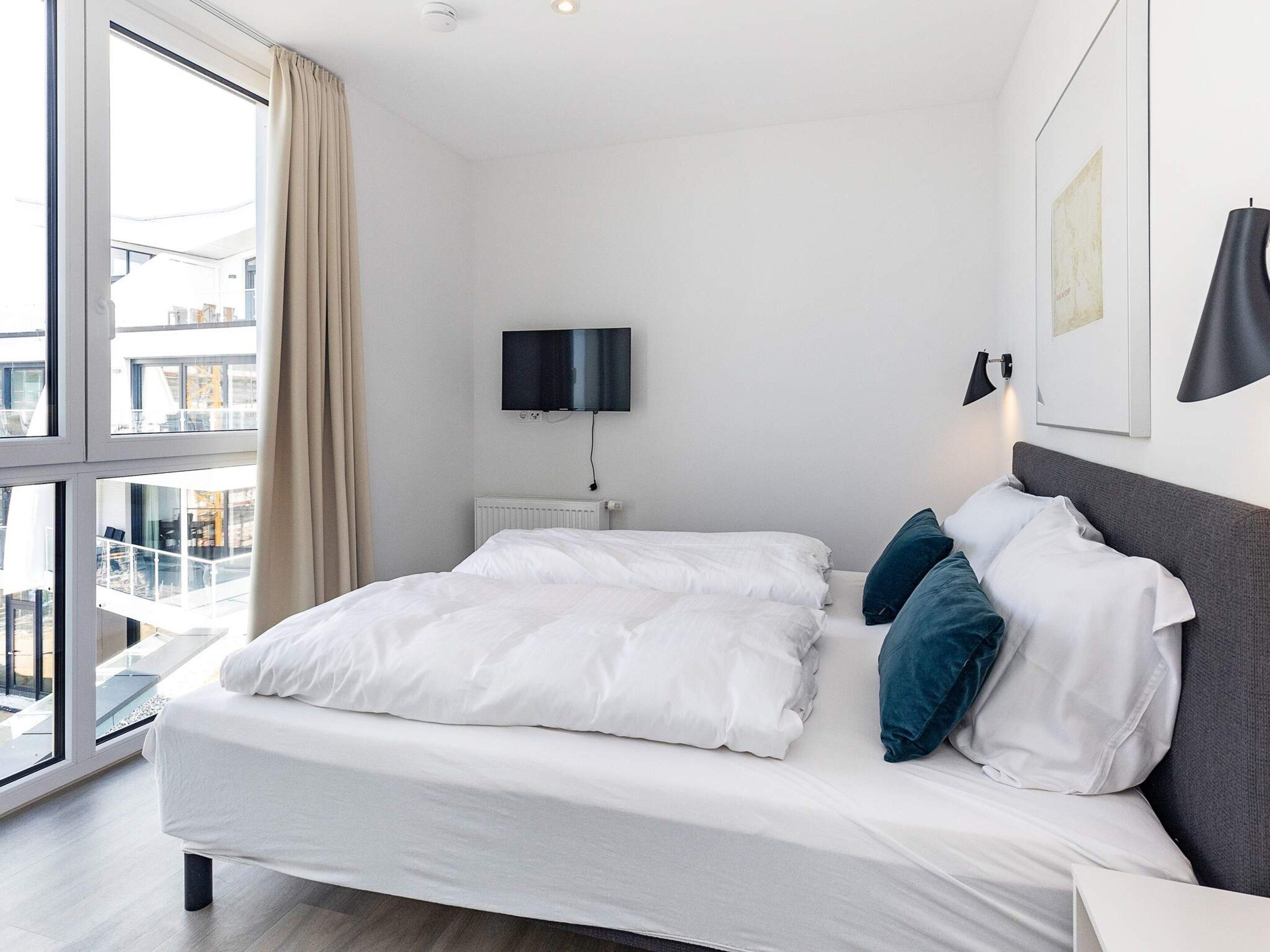 A modern hotel room with a large bed, a flat-screen TV mounted on the wall, and a view of the city through large windows.
