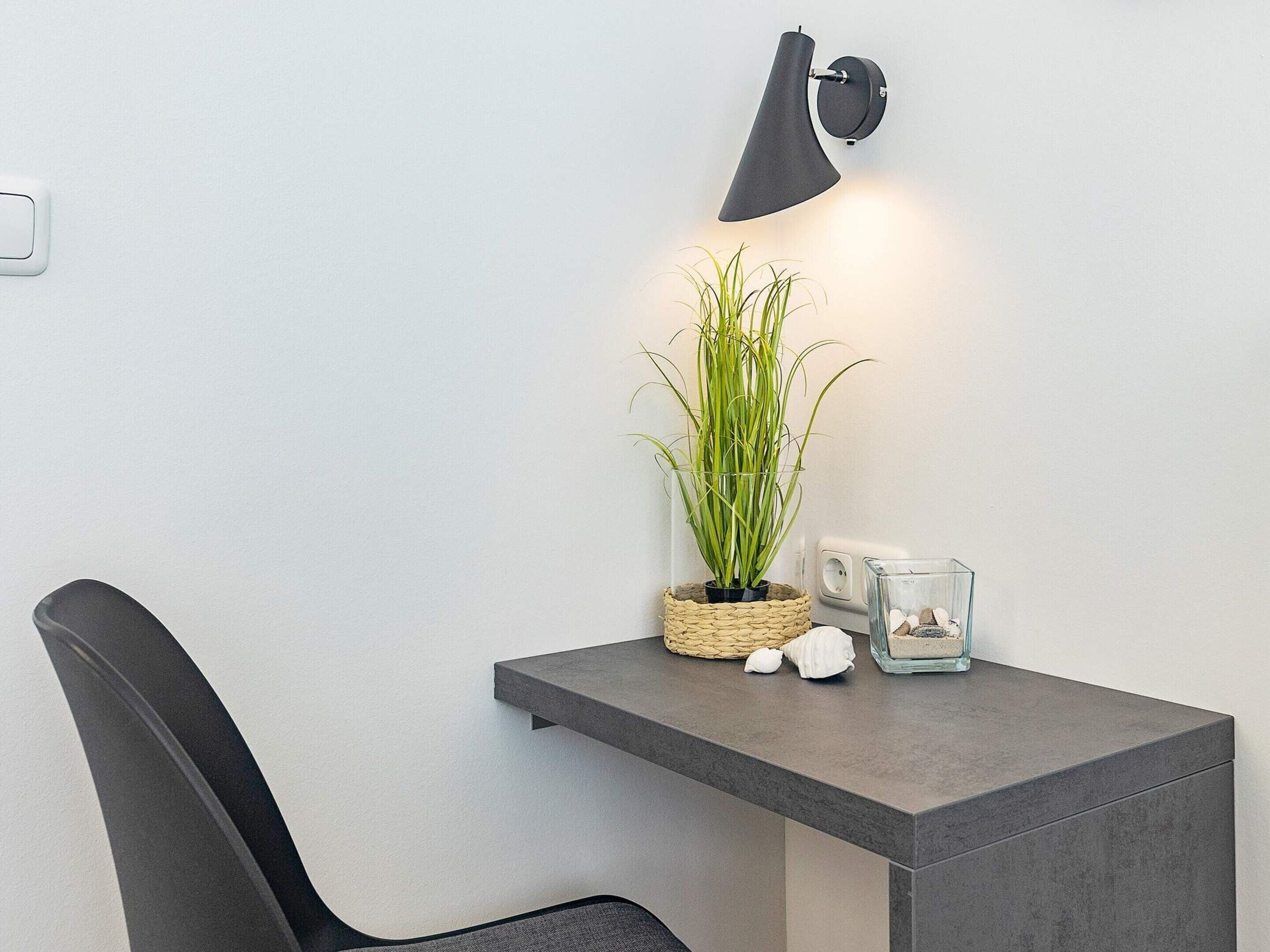 Quarto moderno com escrivaninha cinza, cadeira preta, planta em vaso e luminária de parede.