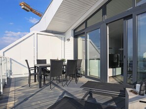 Um deck externo com mesa e cadeiras, um banco e uma porta de vidro que dá para um espaço interno.