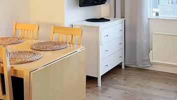 Quarto com mesa de jantar de madeira, cadeiras, cômoda branca e televisão na parede.