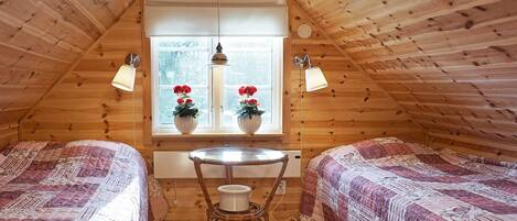 A cozy attic bedroom with two beds, a small table, and a window with potted plants.