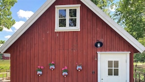 Un edificio de madera rojo con puerta y ventana blancas, rodeado de helechos y plantas en macetas.