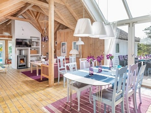 A cozy, well-lit room with a wooden ceiling, a fireplace, a dining table with chairs, and a large window offering a view of the outdoors.