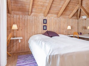 A bedroom with a wooden ceiling, a bed with white bedding, two bedside lamps, a small desk with a chair, and a purple pillow.