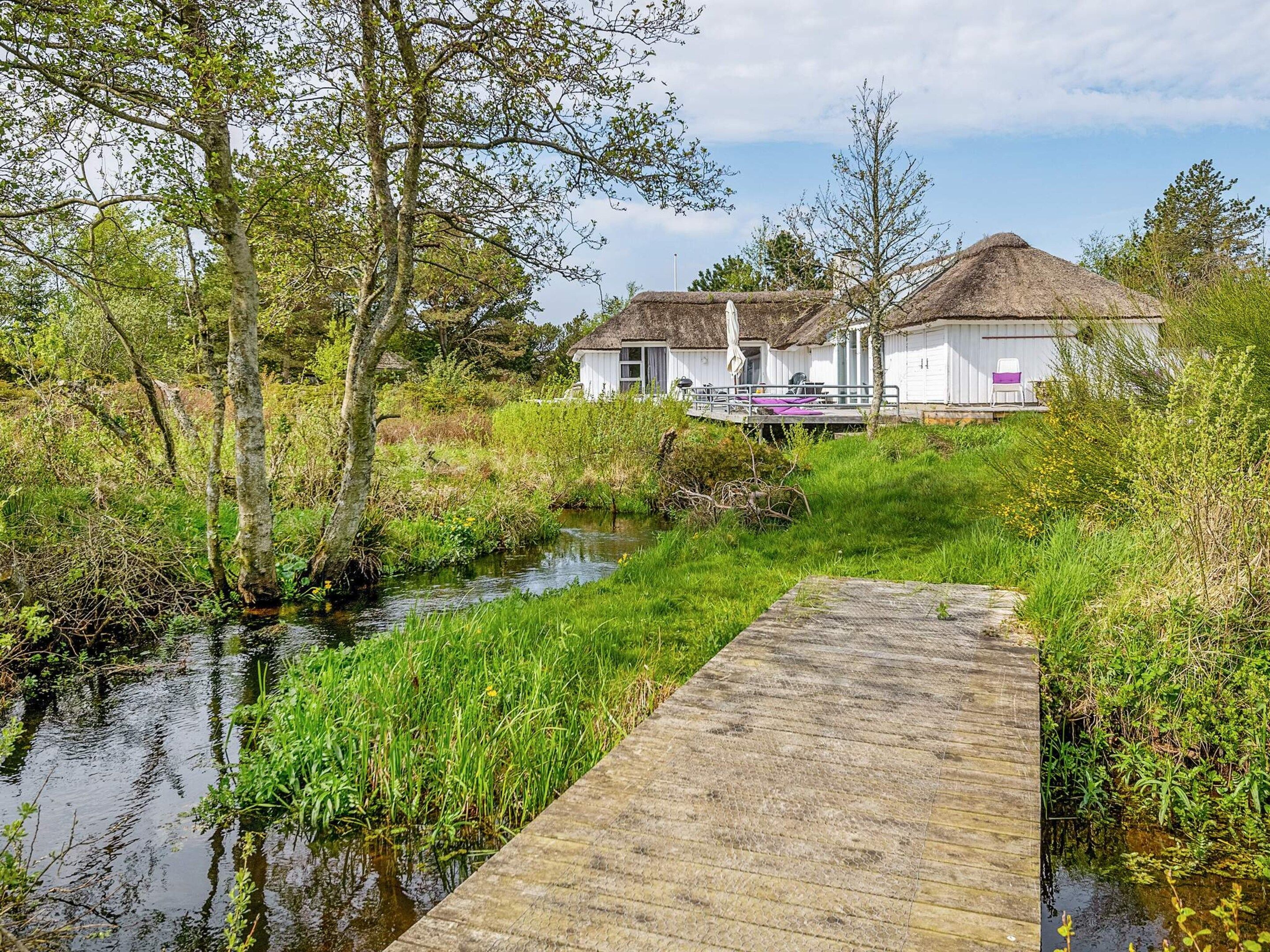 A wooden dock leading to a white building with a thatched roof, surrounded by greenery and a calm water body.