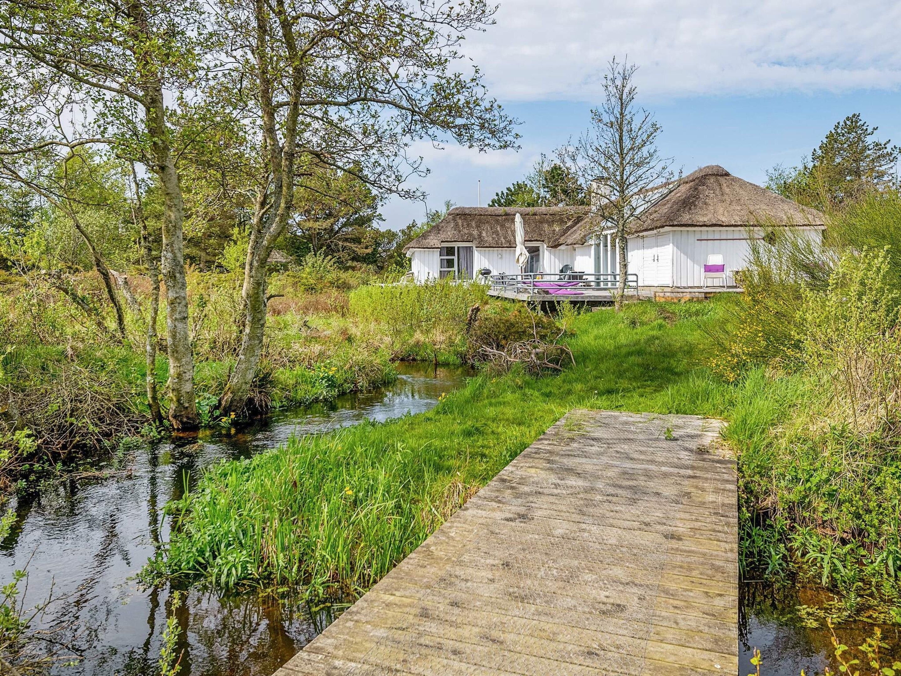 A wooden dock leading to a row of white buildings with thatched roofs, surrounded by greenery and a calm water body.
