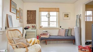 Una sala acogedora con un sillón de mimbre, un sofá con cojines estampados, una ventana adornada con un adorno y una pared con obras de arte enmarcadas.