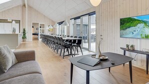 A spacious living area with a long dining table, modern furniture, and a large TV screen displaying a scenic view. - 5 Star Holiday Home in Spottrup (Spøttrup)
