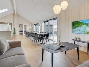 A spacious living area with a long dining table, modern furniture, and a large TV screen displaying a scenic view. - 5 Star Holiday Home in Spottrup (Spøttrup)
