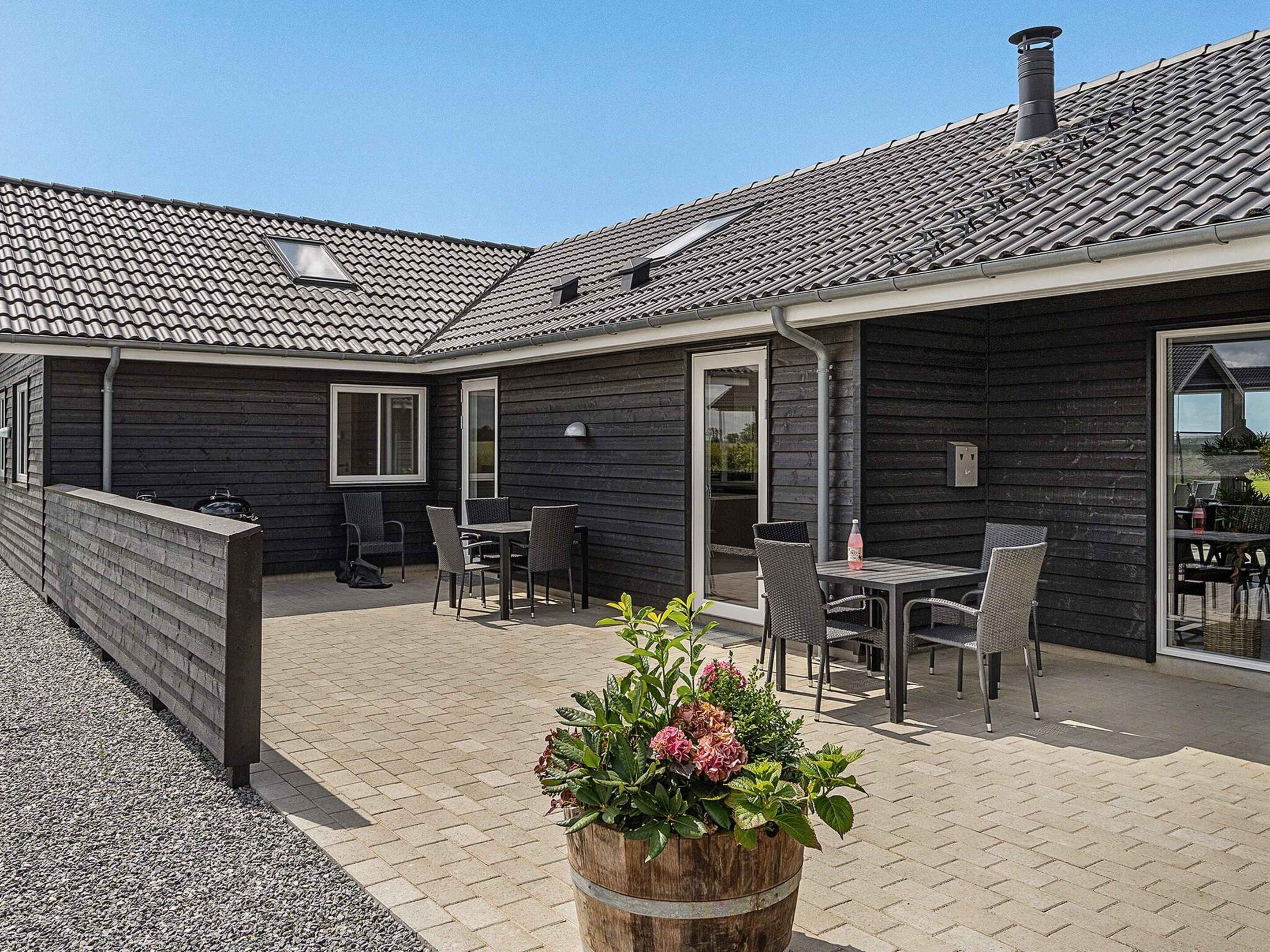 Un patio techado al aire libre con una mesa y sillas, una jardinera de madera con flores, y un edificio con tejado de baldosas.
