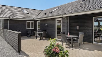 Un patio techado al aire libre con una mesa y sillas, una jardinera de madera con flores, y un edificio con tejado de baldosas.