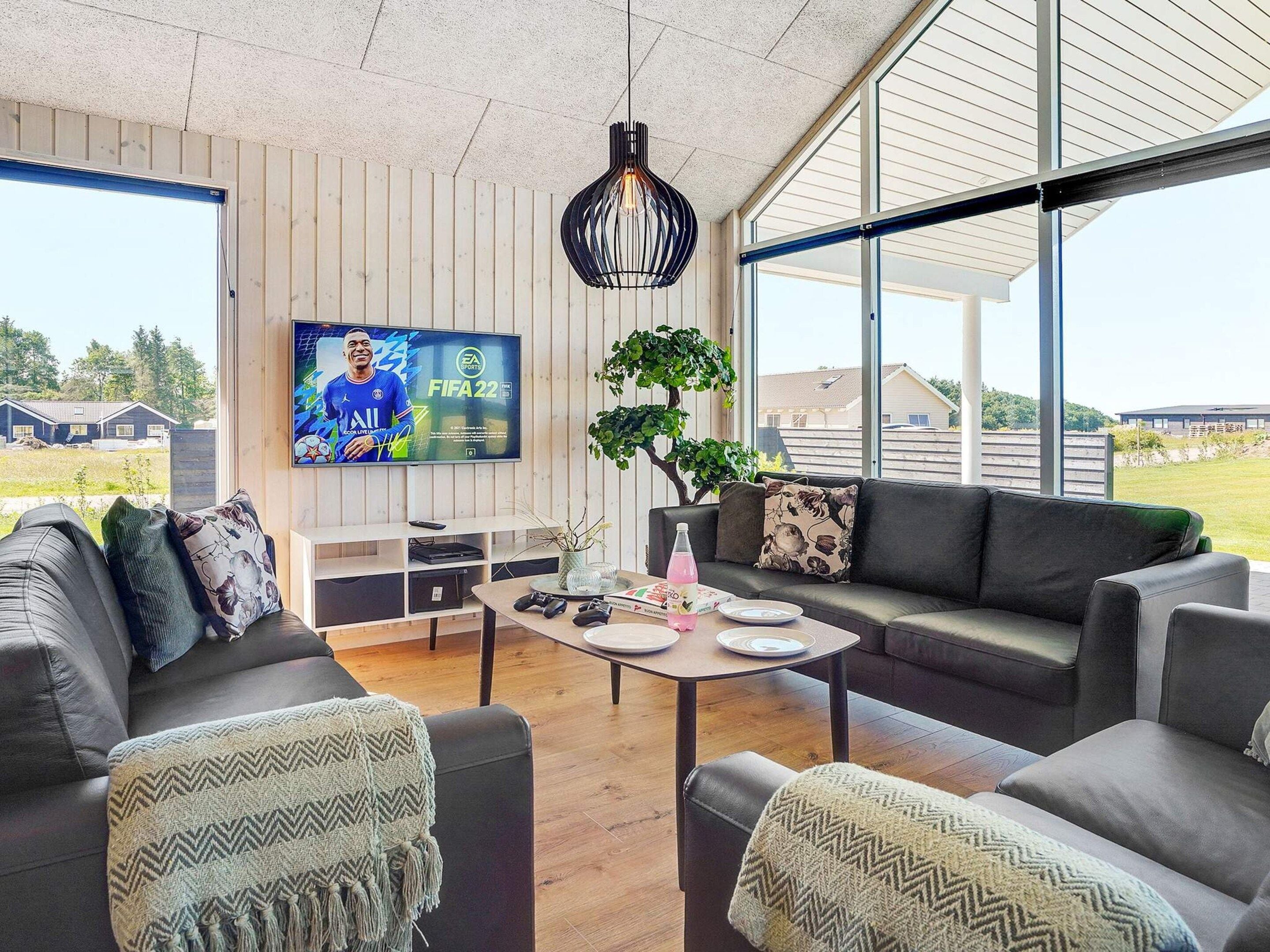 Una sala de estar moderna con un televisor mostrando FIFA22, un conjunto de sofás, una mesa de centro y vistas al exterior.