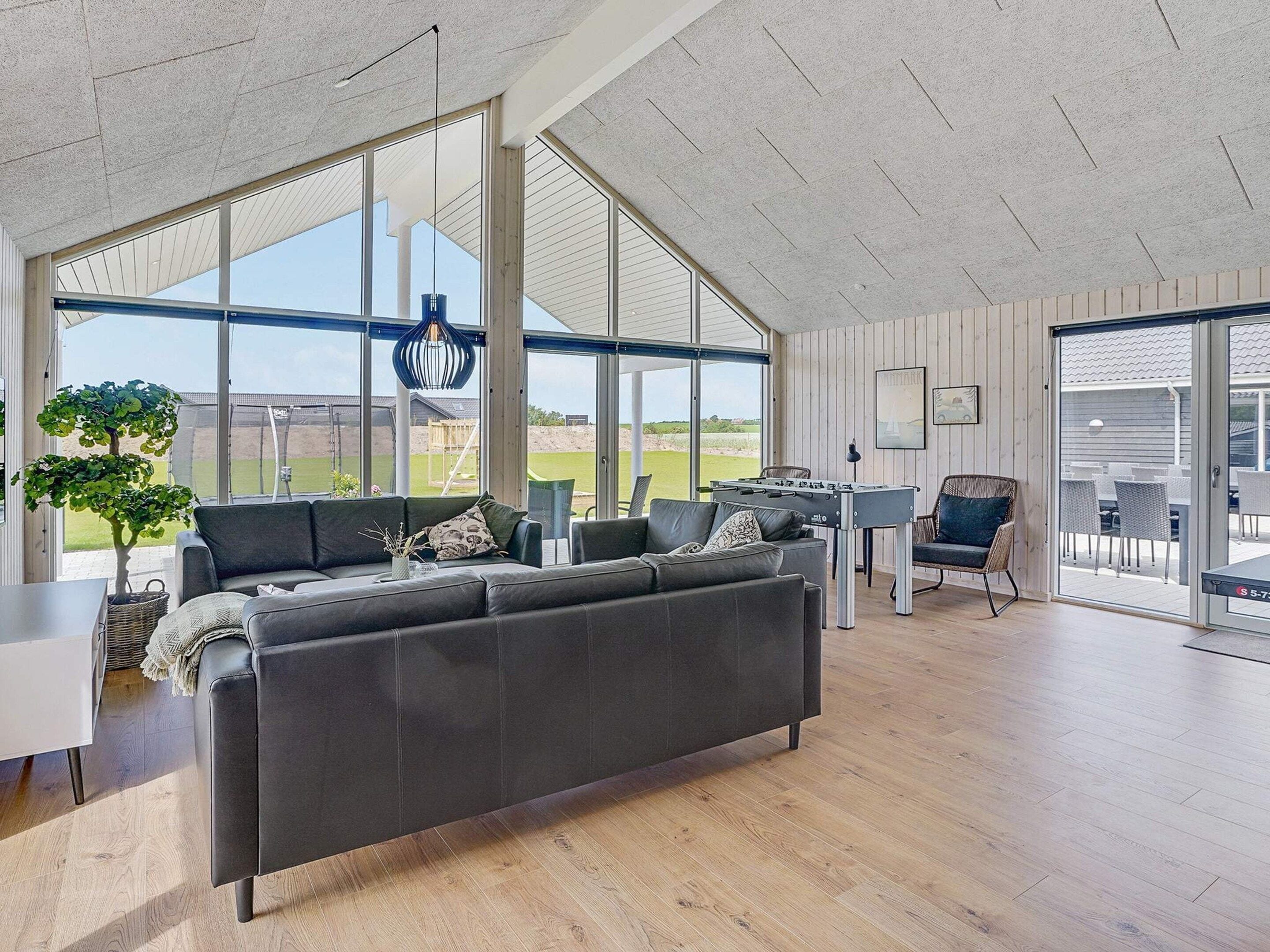 Un salón amplio con un sofá grande, una mesa de futbolín y vistas al exterior a través de amplias ventanas.