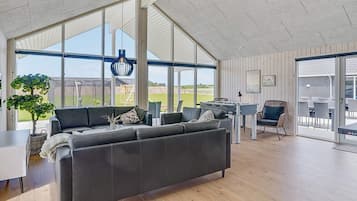 Un salón amplio con un sofá grande, una mesa de futbolín y vistas al exterior a través de amplias ventanas.