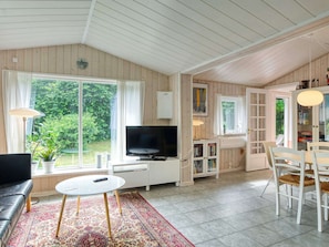 A room with a TV, a dining table, chairs, a bookshelf, and a window with curtains.