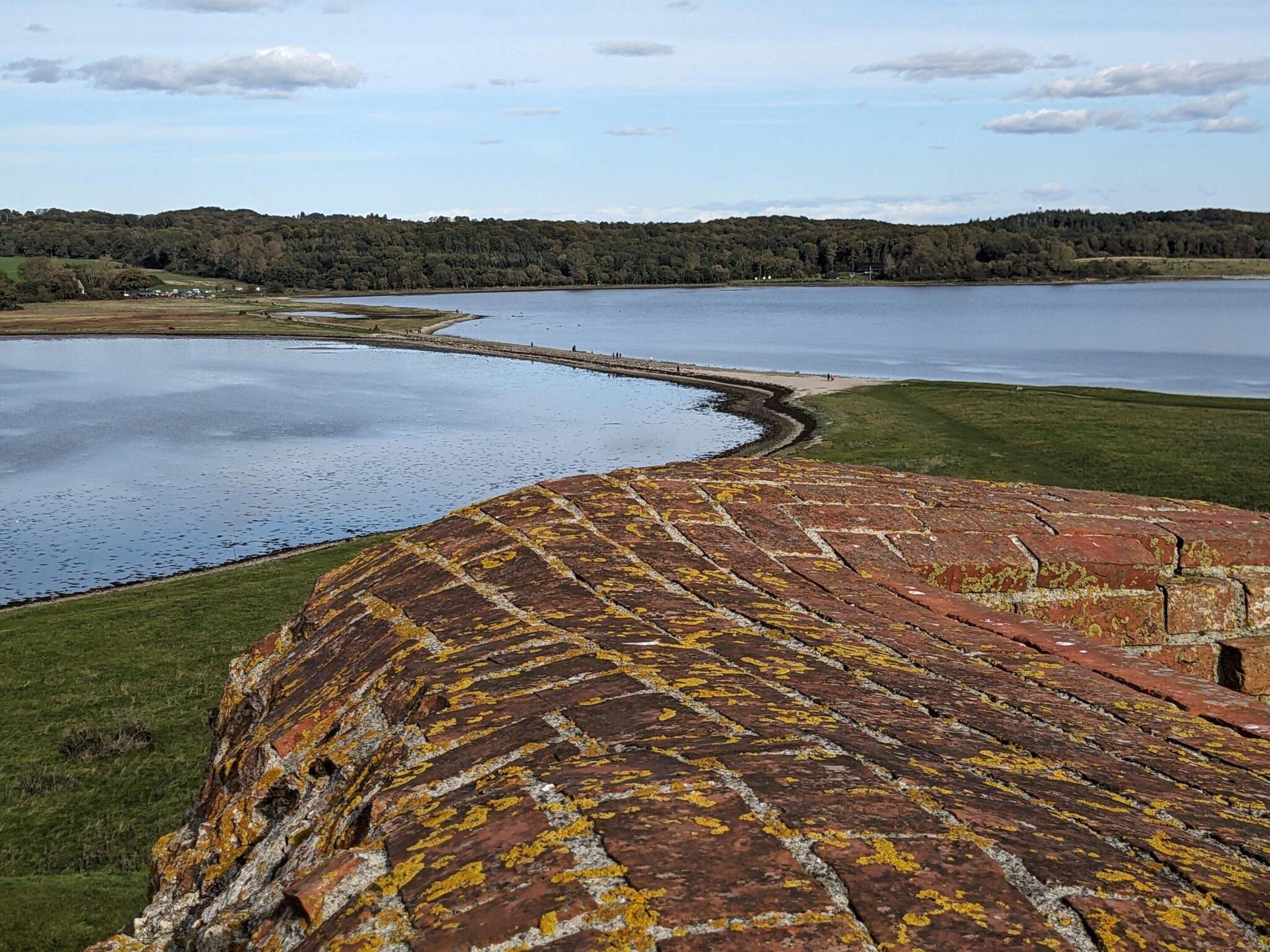 Paisaje costero con una estructura curva de ladrillo, una masa de agua y una colina boscosa al fondo.