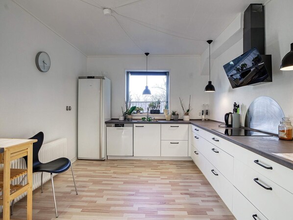A modern kitchen with white cabinets, a built-in dishwasher, a window with plants, and a dining area with a chair and table.