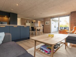 A modern living room with a sofa, a coffee table with books, a dining area with chairs, and a kitchen area with cabinets.