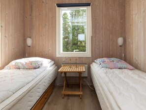 A room with two beds, a window with a view of greenery, and a small wooden table with a lamp.