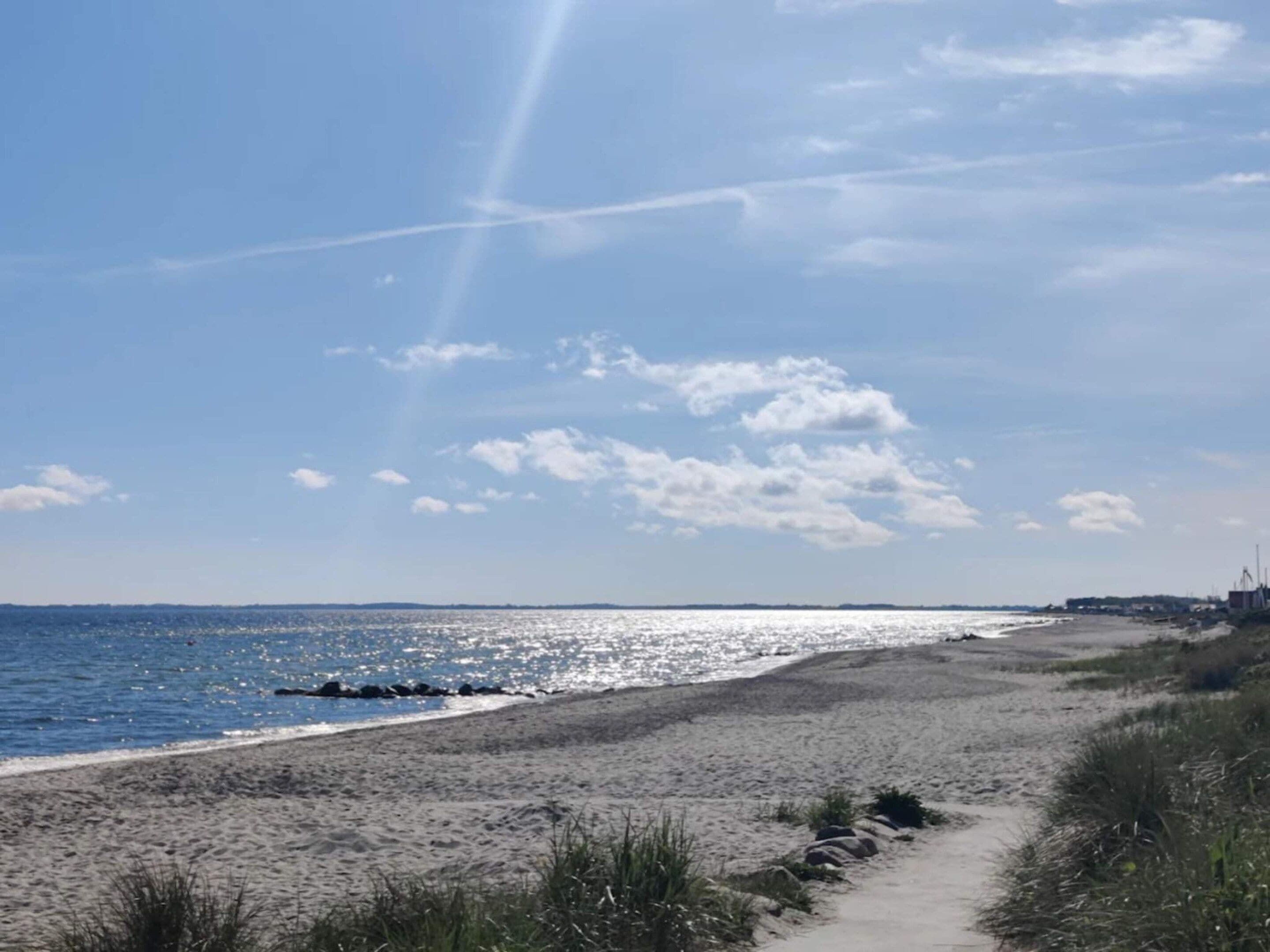 A beach path leading to the ocean with a clear sky and distant industrial structures.