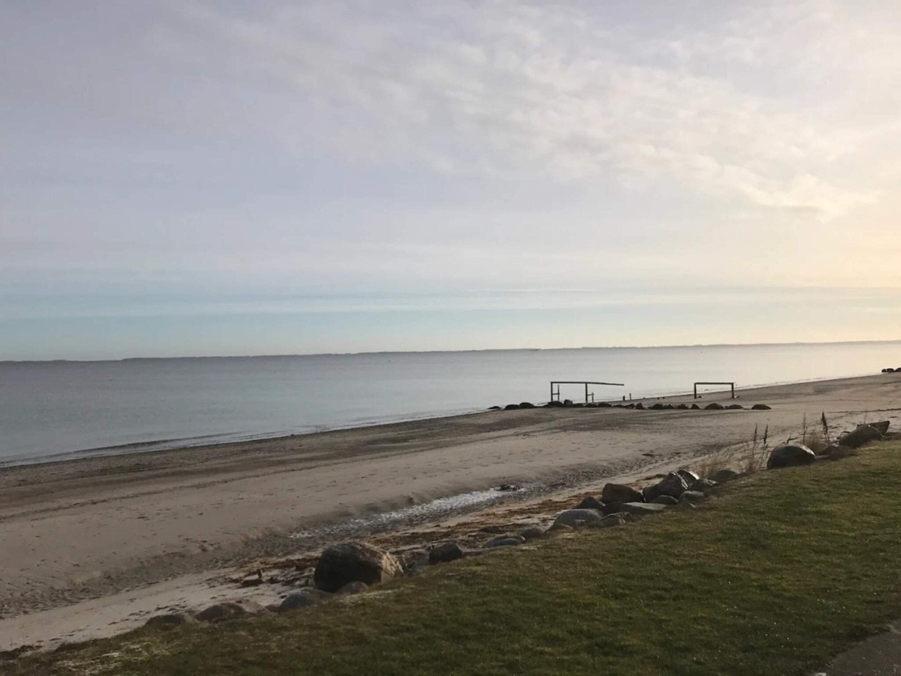 Una playa con una zona cubierta de césped, un sendero de arena y una construcción cerca del agua.