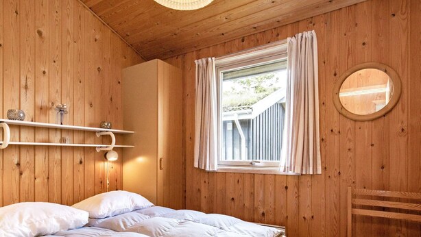 A wooden room with a bed, a chair, a window, and a mirror on the wall. - 6 Person Holiday Home in Blokhus (Blokhus)