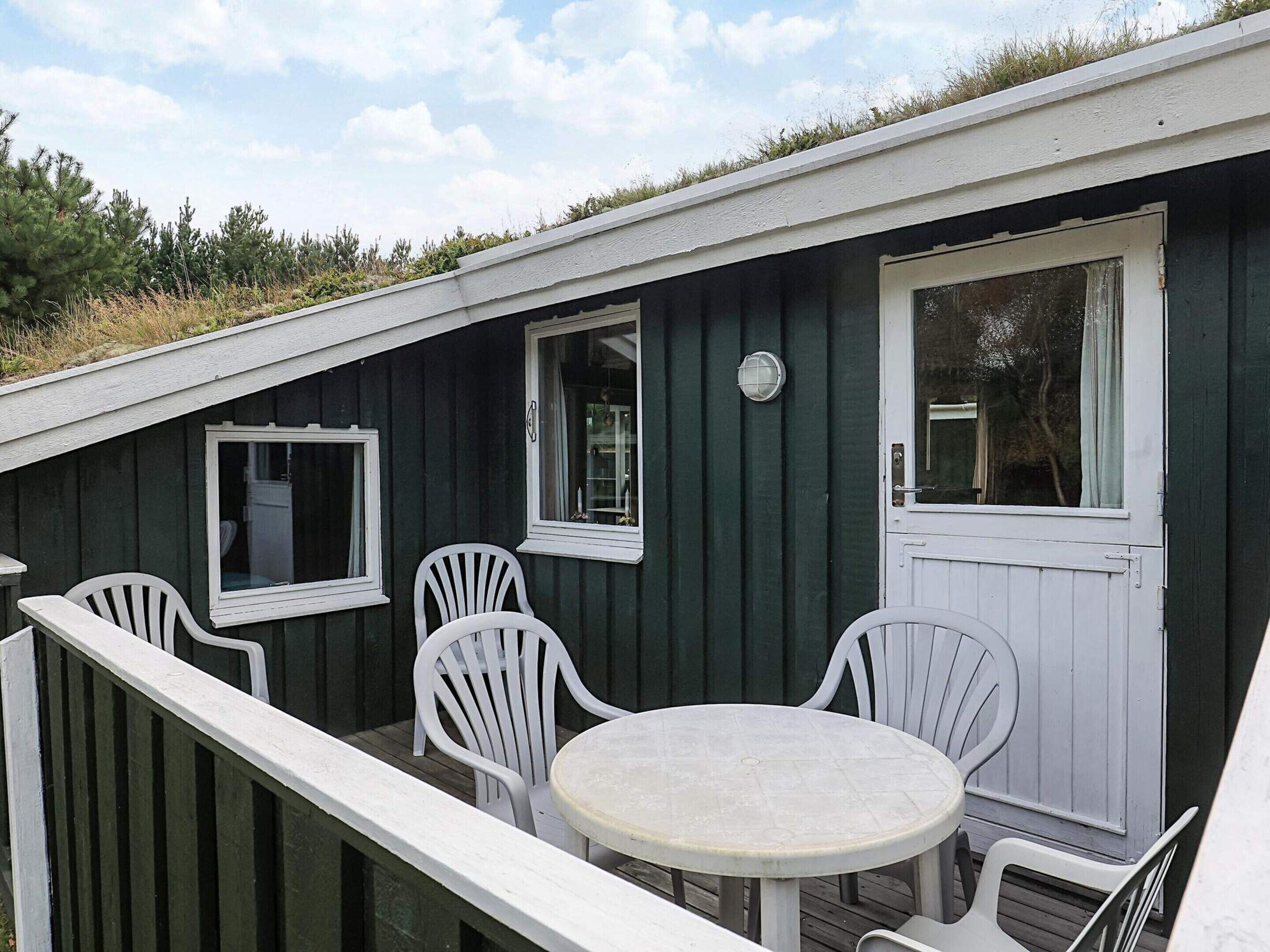 A small, dark green wooden building with white trim, a round table, and four white chairs.