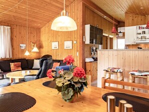 A cozy room with a wooden ceiling and walls, a leather sofa, a dining table with a vase of flowers, and a kitchen area with cabinets and a refrigerator.