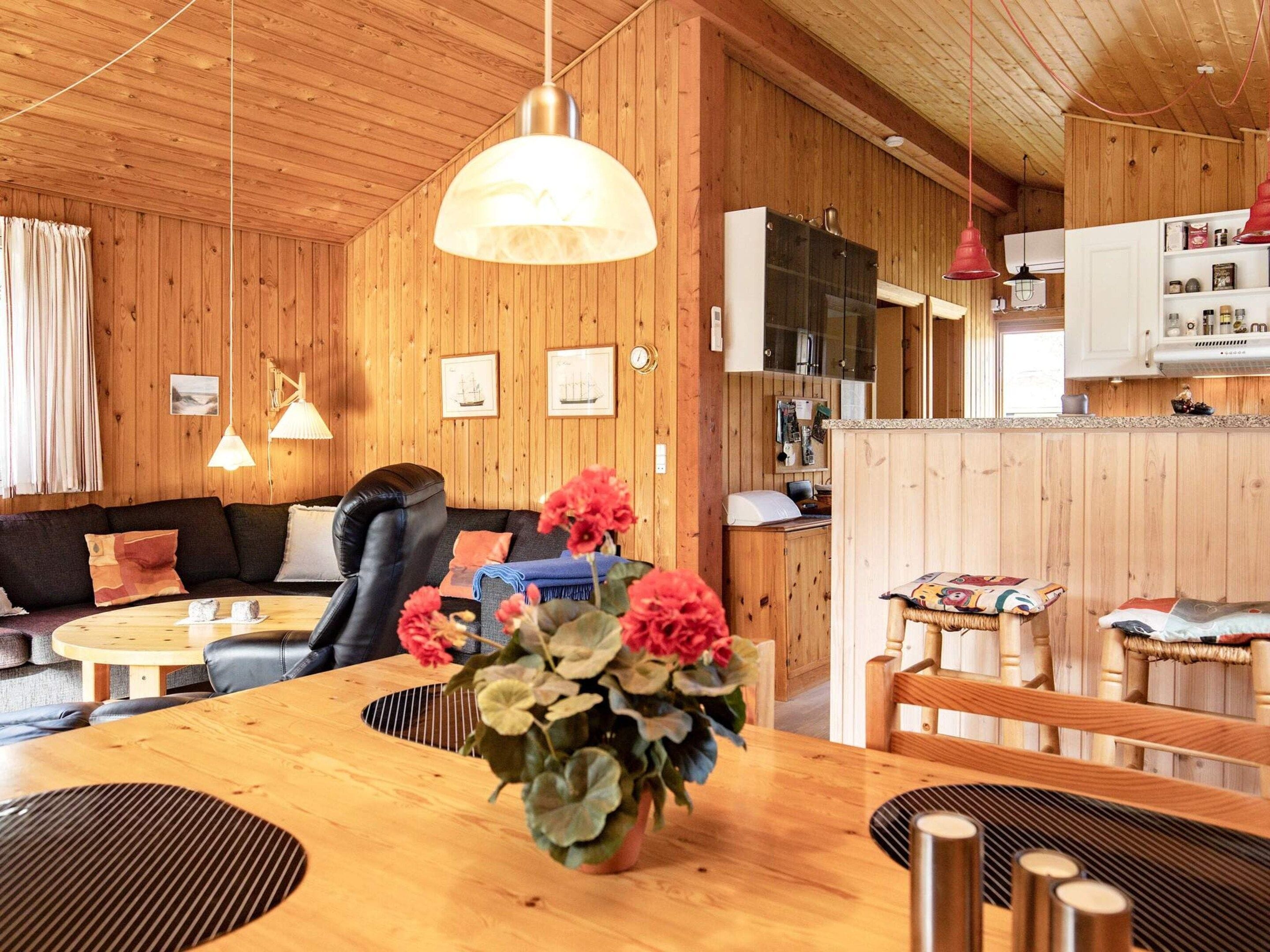 A cozy room with a wooden ceiling, a sofa, a dining table with a vase of flowers, and a kitchen area with cabinets and a refrigerator.