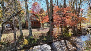 Escape to a serene forest retreat with a cozy cabin, chimney, and porch overlooking a nearby stream.