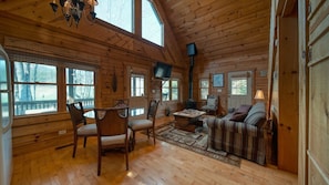 A cozy cabin interior with a wooden floor, a dining table with chairs, a sofa, a television, and large windows offering a view of the outdoors. - Creekside Fish Cabin (Hiawassee)