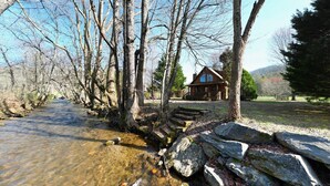 A cabin by a clear stream with a stone bridge and a wooden house in the background. - Creekside Fish Cabin (Hiawassee)