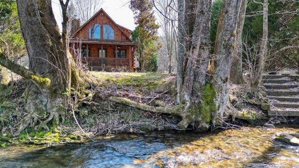 A cabin with a wooden exterior, surrounded by trees and a stream. - Creekside Fish Cabin (Hiawassee)