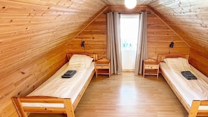 Een kleine zolderkamer met houten lambrisering, twee eenpersoonsbedden, een raam met gordijnen en een houten vloer.