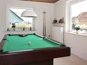 A pool table with a green felt surface, a white overhead light, and a window with a view of a building and greenery outside.
