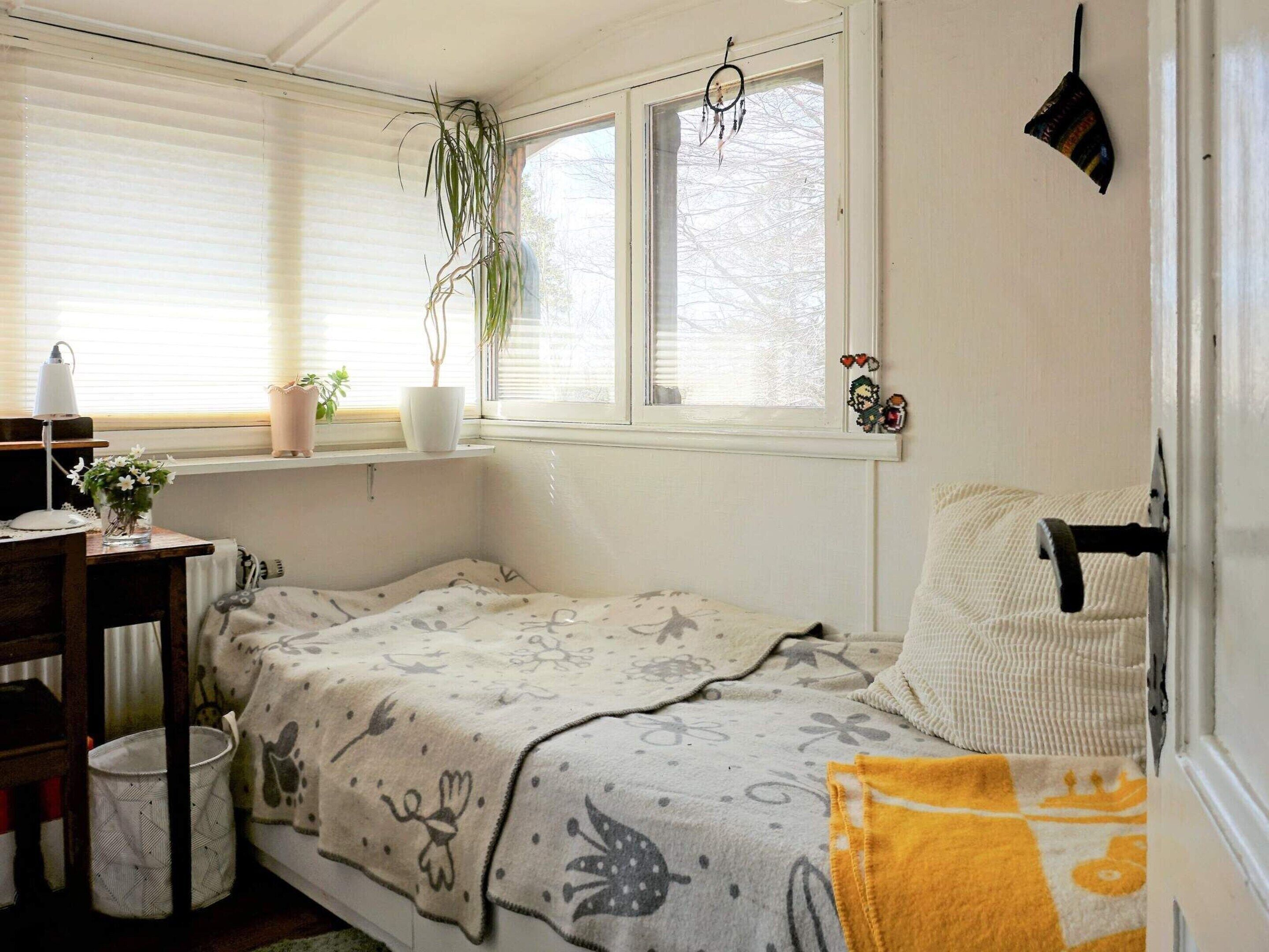 Ett litet, mysigt sovrum med en enkel säng, ett skrivbord med en lampa, ett fönster med persienner och en upphängd drömfångare.